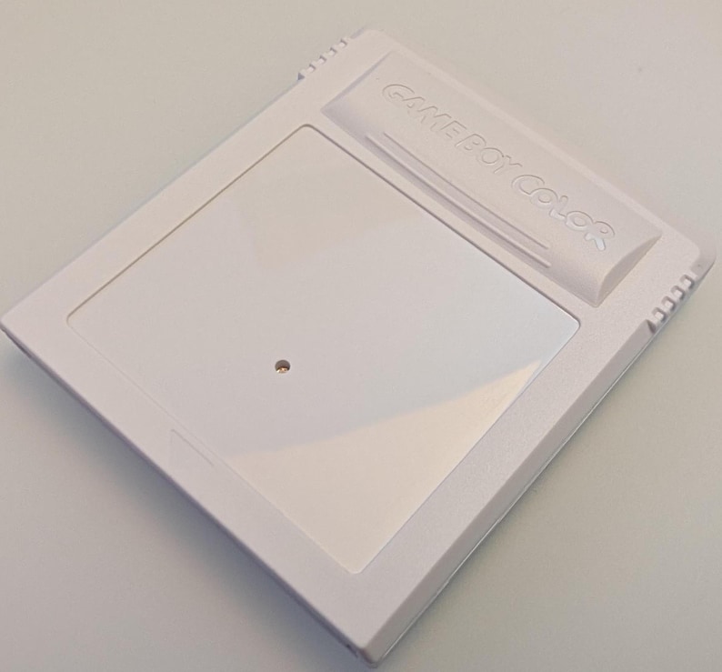 May include: A white Game Boy Color cartridge. The cartridge has a rectangular design with a recessed area and the words "GAME BOY COLOR" embossed on the top. A small, circular indentation is visible on the front.