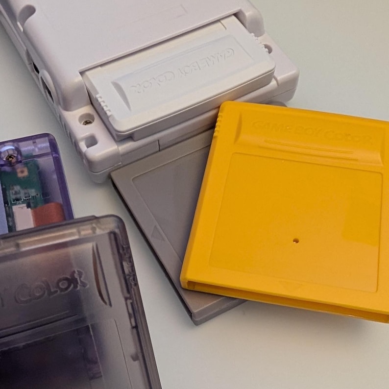May include: A white Game Boy Color console with an open cartridge slot, next to several game cartridges. One cartridge is yellow, another is gray, and a third is purple. The text "GAME BOY COLOR" is visible on the cartridges.