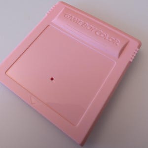May include: A light pink Game Boy Color cartridge. The cartridge has the words "GAME BOY COLOR" embossed on the top. The cartridge has a rectangular shape with a small hole and a triangle symbol.