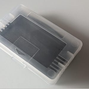 May include: A clear, rectangular plastic box with a hinged lid. The box contains a dark gray, rectangular object. The box is slightly translucent, allowing a glimpse of the contents within. The box is on a light gray surface.