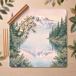 May include: A square mahjong mat featuring a printed landscape of a snow-capped mountain reflected in a calm lake, surrounded by pine trees and blueberry bushes. A collection of ivory-colored mahjong tiles and two wooden tile racks are placed on top.