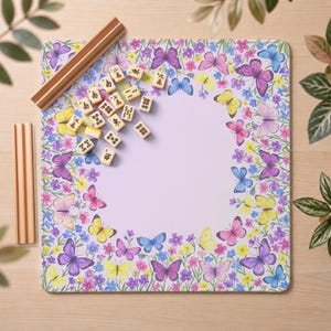 May include: A square lavender game mat featuring a border of colorful butterflies and small flowers, accompanied by a set of small rectangular mahjong tiles and two wooden tile racks positioned on the left side of the mat.