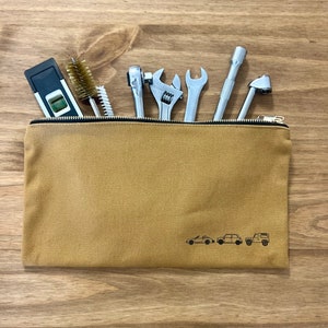 May include: A tan canvas tool bag with a zipper closure. The bag has three small cars printed on the front. The bag is filled with tools, including a spirit level, a wrench, a socket wrench, a brush, and a tyre gauge.