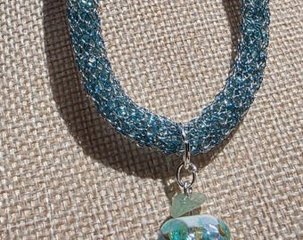 May include: A blue mesh necklace with a pendant. The necklace is made of a woven, light blue material. The pendant is a colorful, round bead with a turquoise base and a small green stone accent.