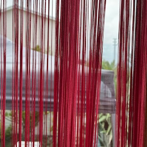 Può includere: Tende a corda rosse appese a una finestra, con una vista sfocata di un edificio e vegetazione all'esterno.