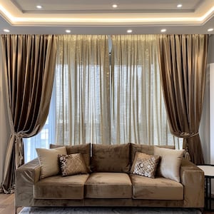 May include: A brown velvet sofa with three cushions and a patterned throw pillow. The sofa is in front of a window with brown velvet curtains and sheer gold curtains.