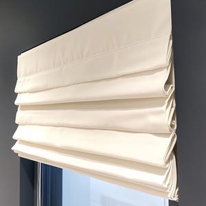 May include: White roman shade window blind with a pleated design. The shade is fully extended and is mounted on a wall.