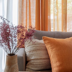 May include: A close-up of a gray sofa with two throw pillows. The sofa is in front of a window with orange sheer curtains. A vase with dried flowers sits on a wooden table in the foreground.