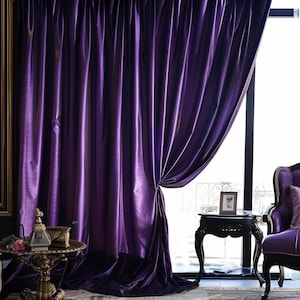 May include: Purple velvet curtains with a satin sheen, hanging in a room with a side table and a chair. The curtains are drawn back to reveal a window with a view of the outdoors.