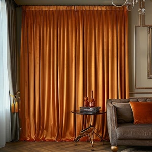May include: A pair of orange velvet curtains hang in a living room with a brown leather sofa and a small side table with a vase and books.