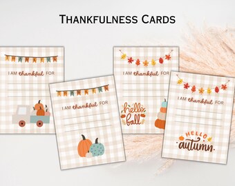 May include: Four printable Thanksgiving cards with a gingham background. The cards feature autumn-themed illustrations, including pumpkins, leaves, and a truck carrying pumpkins. Each card has the text "I am thankful for" and a blank space for writing.