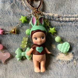 Puede incluir: Un llavero de mu&ntilde;eca verde y marr&oacute;n con un coraz&oacute;n que dice "YOU" en la parte delantera. La mu&ntilde;eca lleva un gorro de conejito verde y tiene orejas de conejito rosas. El llavero est&aacute; decorado con estrellas verdes, corazones y lazos.