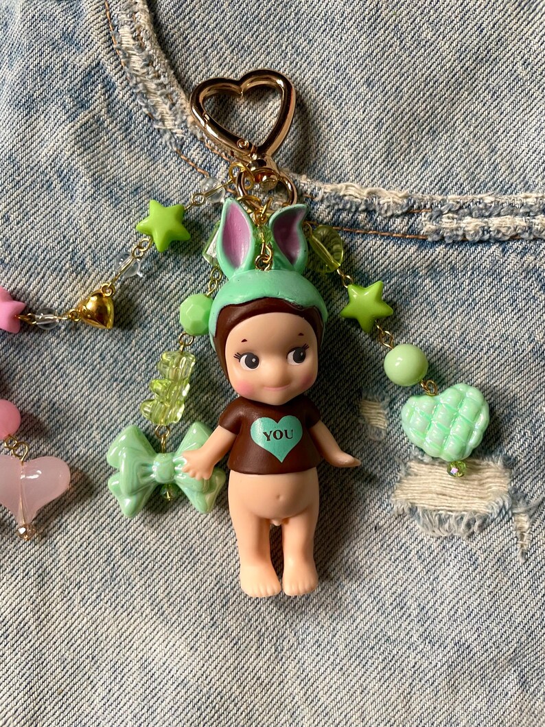 Puede incluir: Un llavero de mu&ntilde;eca verde y marr&oacute;n con un coraz&oacute;n que dice "YOU" en la parte delantera. La mu&ntilde;eca lleva un gorro de conejito verde y tiene orejas de conejito rosas. El llavero est&aacute; decorado con estrellas verdes, corazones y lazos.