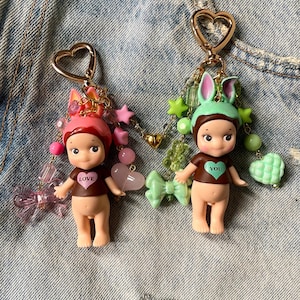 Puede incluir: Dos peque&ntilde;os llaveros de mu&ntilde;eca con anillos de coraz&oacute;n dorados. Las mu&ntilde;ecas est&aacute;n desnudas con camisas marrones que dicen "LOVE" y "YOU". Las mu&ntilde;ecas est&aacute;n adornadas con cuentas rosas, verdes y doradas, estrellas y lazos.