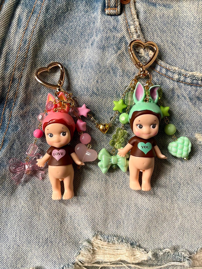 Puede incluir: Dos peque&ntilde;os llaveros de mu&ntilde;eca con anillos de coraz&oacute;n dorados. Las mu&ntilde;ecas est&aacute;n desnudas con camisas marrones que dicen "LOVE" y "YOU". Las mu&ntilde;ecas est&aacute;n adornadas con cuentas rosas, verdes y doradas, estrellas y lazos.