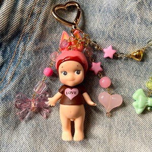 Puede incluir: Un llavero de mu&ntilde;eca rosa y marr&oacute;n con un dije en forma de coraz&oacute;n y la palabra "LOVE" en la camiseta de la mu&ntilde;eca. El llavero est&aacute; decorado con lazos rosas y verdes, estrellas y corazones.