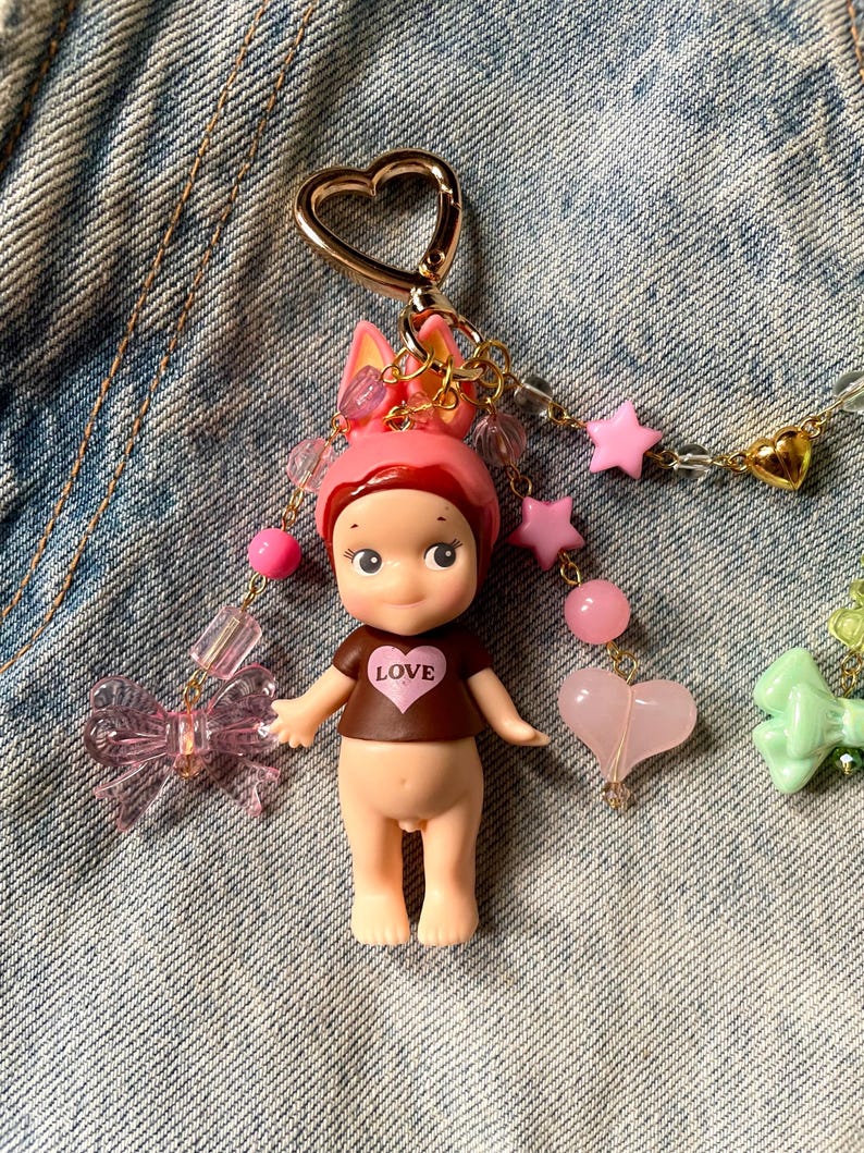 Puede incluir: Un llavero de mu&ntilde;eca rosa y marr&oacute;n con un dije en forma de coraz&oacute;n y la palabra "LOVE" en la camiseta de la mu&ntilde;eca. El llavero est&aacute; decorado con lazos rosas y verdes, estrellas y corazones.