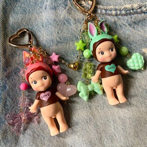 Puede incluir: Dos llaveros con dijes que representan una peque&ntilde;a mu&ntilde;eca con cuerpo marr&oacute;n y una camisa marr&oacute;n con un coraz&oacute;n. La mu&ntilde;eca de la izquierda lleva un gorro de conejito rosa y dijes rosas y verdes. La mu&ntilde;eca de la derecha lleva un gorro de conejito verde y dijes verdes y dorados. Ambas mu&ntilde;ecas tienen un coraz&oacute;n en su camisa, uno dice "LOVE" y el otro dice "YOU".