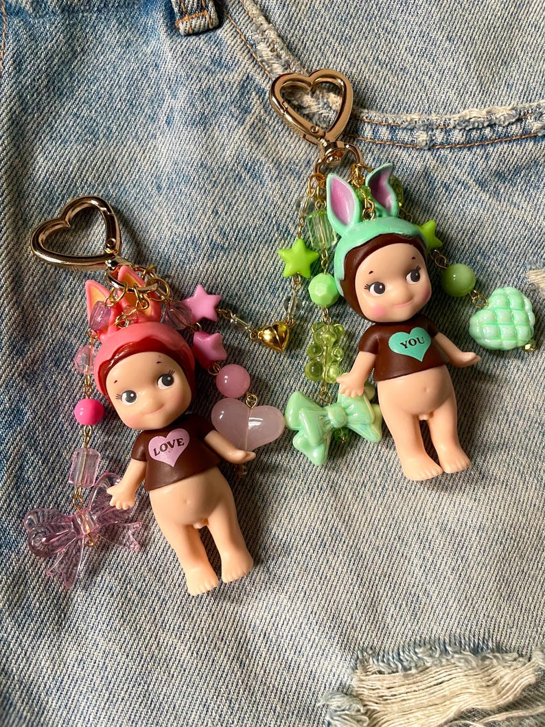 Puede incluir: Dos llaveros con dijes que representan una peque&ntilde;a mu&ntilde;eca con cuerpo marr&oacute;n y una camisa marr&oacute;n con un coraz&oacute;n. La mu&ntilde;eca de la izquierda lleva un gorro de conejito rosa y dijes rosas y verdes. La mu&ntilde;eca de la derecha lleva un gorro de conejito verde y dijes verdes y dorados. Ambas mu&ntilde;ecas tienen un coraz&oacute;n en su camisa, uno dice "LOVE" y el otro dice "YOU".