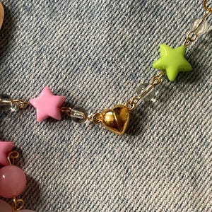 Puede incluir: Un collar de cadena dorada con cuentas en forma de estrella rosa y verde y un dije de coraz&oacute;n dorado.