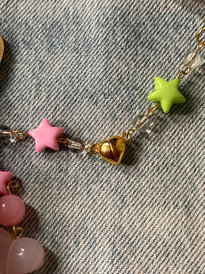Puede incluir: Un collar de cadena dorada con cuentas en forma de estrella rosa y verde y un dije de coraz&oacute;n dorado.