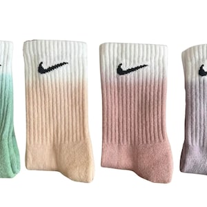 May include: Four pairs of Nike socks in a row, each with a different color gradient. The socks are white at the top, fading into green, peach, pink, and purple. A black Nike swoosh logo is embroidered on each sock.