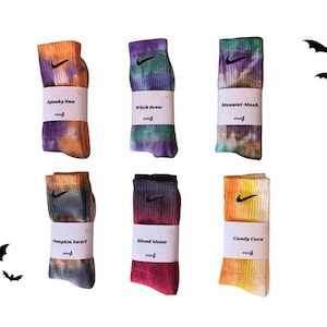 May include: Six pairs of Nike socks with tie-dye patterns and the Nike swoosh logo. Each pair is wrapped with a label featuring a Halloween-themed name: Spooky Boo, Witch Brew, Monster Mash, Pumpkin Swirl, Blood Moon, and Candy Corn. Black bat silhouettes are in the background.