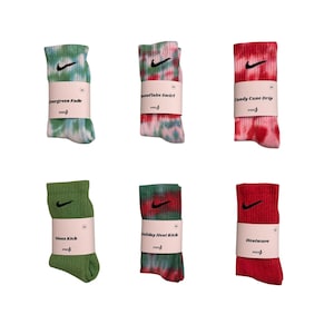 May include: Six pairs of Nike socks in various colors and patterns. The socks include designs like tie-dye, solid green, and red. Each pair is wrapped with a paper band featuring the Nike swoosh logo and a product name such as "Evergreen Fade" and "Heatwave."