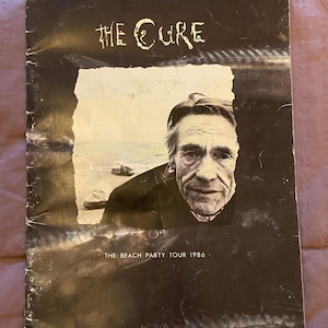 May include: A rectangular concert tour program for The Cure, featuring a black and white portrait of a person against a beach background. The text reads THE CURE at the top and THE BEACH PARTY TOUR 1986 at the bottom. The cover has a dark, textured border.