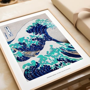以下が含まれることがあります： 葛飾北斎の「神奈川沖浪裏」の額装プリント。このアートワークは、青と水色の色合いで描かれた大きく様式化された波と、背景に雪をかぶった山が特徴です。プリントは明るい木製のフレームに入っています。