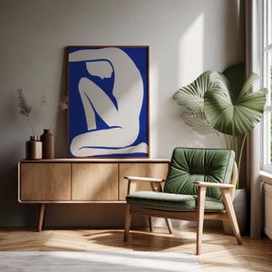 Puede incluir: Una silueta blanca de una persona sentada con las piernas cruzadas sobre un fondo azul. La obra de arte está enmarcada y colgada en una pared sobre un armario de madera con un sillón verde delante.