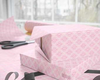 May include: Two gift boxes wrapped in pink patterned paper with a matching roll of paper in the background. Black scissors and a black ribbon are also visible. The wrapping paper has a repeating floral design.