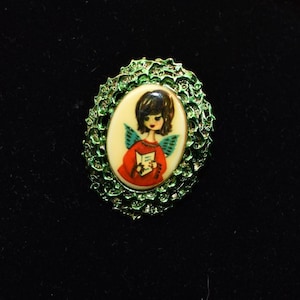May include: Vintage oval brooch with a green filigree frame. The brooch features a painted image of an angel in a red dress holding a book, with black hair and blue wings, set against a cream background.