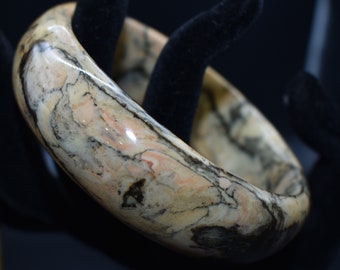 May include: A marbled bangle bracelet with a polished finish. The bracelet features a mix of beige, peach, and black tones, creating a natural stone appearance. The thick, rounded design suggests a statement piece of jewelry.