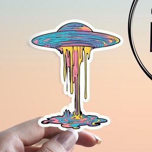 May include: A colorful sticker of a melting UFO with a blue, pink, and yellow dripping design. The sticker is on a hand.