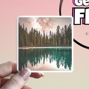Puede incluir: Una pegatina cuadrada que presenta un paisaje sereno con un lago que refleja un bosque de altos árboles de hoja perenne. El cielo es una mezcla de rosa, blanco y gris. La pegatina está sujeta por una mano sobre un fondo rosa. El texto a la derecha dice "BUY FREE".