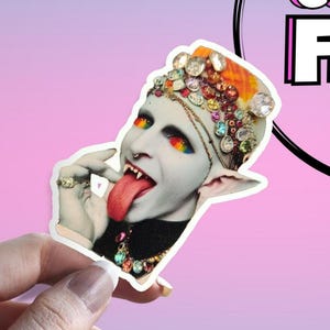 May include: A sticker depicting a stylized image of a person with rainbow eyes and tongue extended. The person wears a jeweled headdress and necklace. The background is a pink and purple gradient.