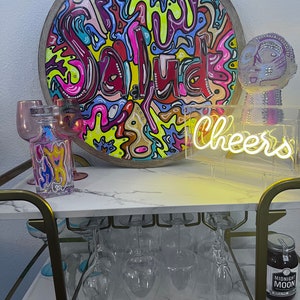 May include: A bar cart with a marble top and gold metal frame. The cart is decorated with a round wooden sign with the word "Salud" written in colourful paint, a neon sign that says "Cheers", and a glass jar filled with a dark liquid. There are also several wine glasses on the cart.