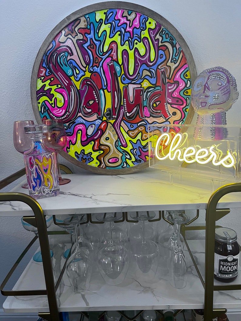 May include: A bar cart with a marble top and gold metal frame. The cart is decorated with a round wooden sign with the word "Salud" written in colourful paint, a neon sign that says "Cheers", and a glass jar filled with a dark liquid. There are also several wine glasses on the cart.