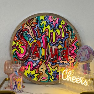 May include: A round wooden tray with a colourful abstract design featuring the word "Salud" in a whimsical font. The tray is surrounded by various decorative items, including a glass decanter, a neon sign that reads "Cheers", and a decorative figurine.