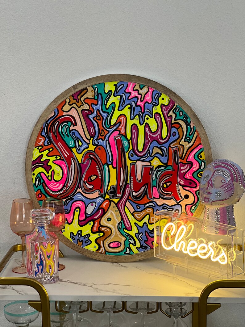 May include: A round wooden tray with a colourful abstract design featuring the word "Salud" in a whimsical font. The tray is surrounded by various decorative items, including a glass decanter, a neon sign that reads "Cheers", and a decorative figurine.