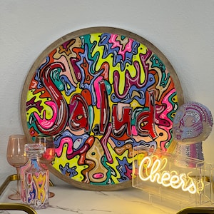 May include: A round wooden tray with a colourful abstract design featuring the word "Salud" in a bright red font. The tray is surrounded by a wooden frame. A neon sign with the word "Cheers" is in the foreground.