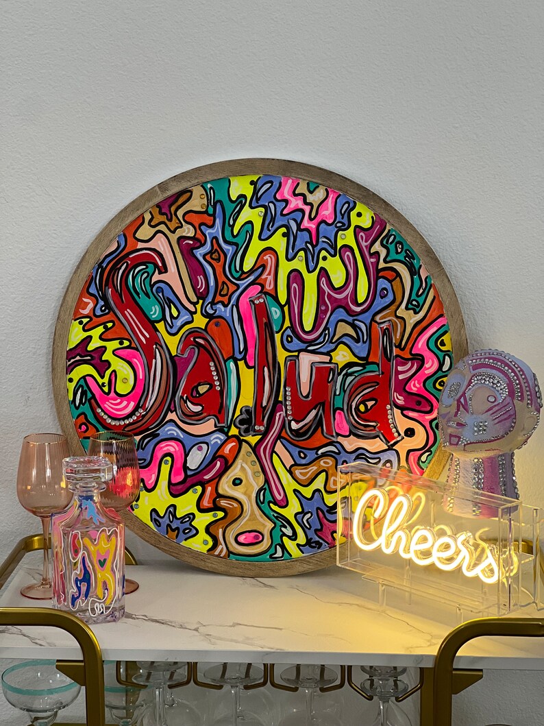 May include: A round wooden tray with a colourful abstract design featuring the word "Salud" in a bright red font. The tray is surrounded by a wooden frame. A neon sign with the word "Cheers" is in the foreground.