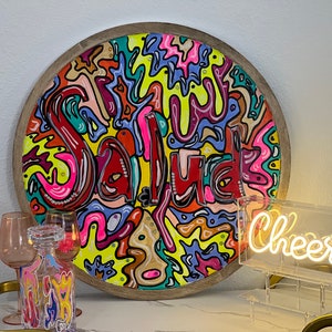May include: A round wooden tray with a colourful abstract design featuring the word "Salud" in red. The tray is on a table with a clear glass neon sign that says "Cheers".