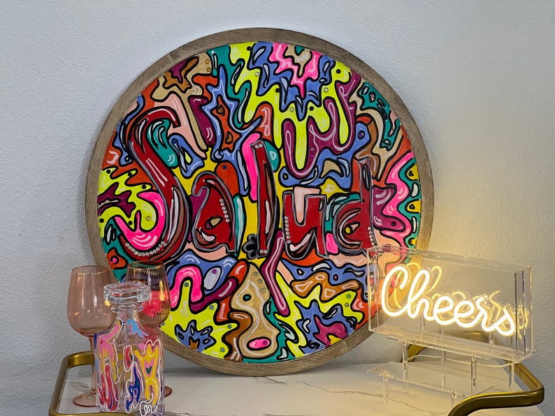 May include: A round wooden tray with a colourful abstract design featuring the word "Salud" in red. The tray is on a table with a clear glass neon sign that says "Cheers".