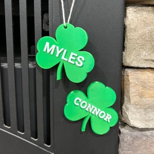 May include: Two green shamrock-shaped ornaments with white lettering. The top ornament says "MYLES" and the bottom ornament says "CONNOR".