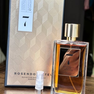 Op de afbeelding: Een flesje van 100 ml Rosendo Mattiu Patchouli Oud Vanille parfum. De fles is van helder glas met een gouden dop en een gouden etiket met de merknaam en de geurbeschrijving. De fles staat op een houten oppervlak naast een klein proefbuisje.