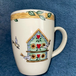 May include: White ceramic mug with a birdhouse design. The birdhouse is green with red heart accents and is surrounded by small birds. The top of the mug has a wood-grain pattern with green leaves.