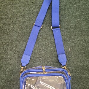 May include: A clear crossbody bag with a blue adjustable strap and blue trim. The bag has a gold zipper and gold-colored hardware. The bag is made of clear plastic, allowing visibility of the contents.