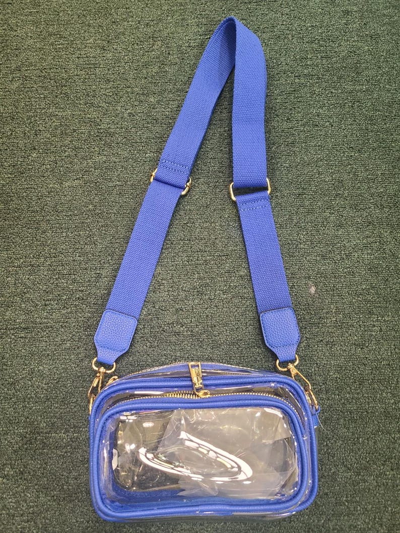 May include: A clear crossbody bag with a blue adjustable strap and blue trim. The bag has a gold zipper and gold-colored hardware. The bag is made of clear plastic, allowing visibility of the contents.